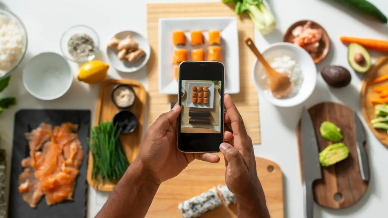 Person photographing homemade sushi preparation with smartphone, ingredients spread on table.