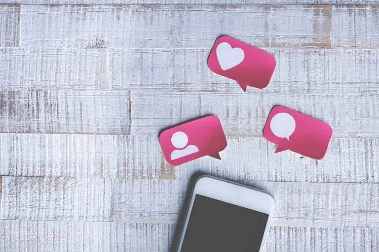 A smartphone with three pink social media icons floating above it, set against a white wooden background. The phone is