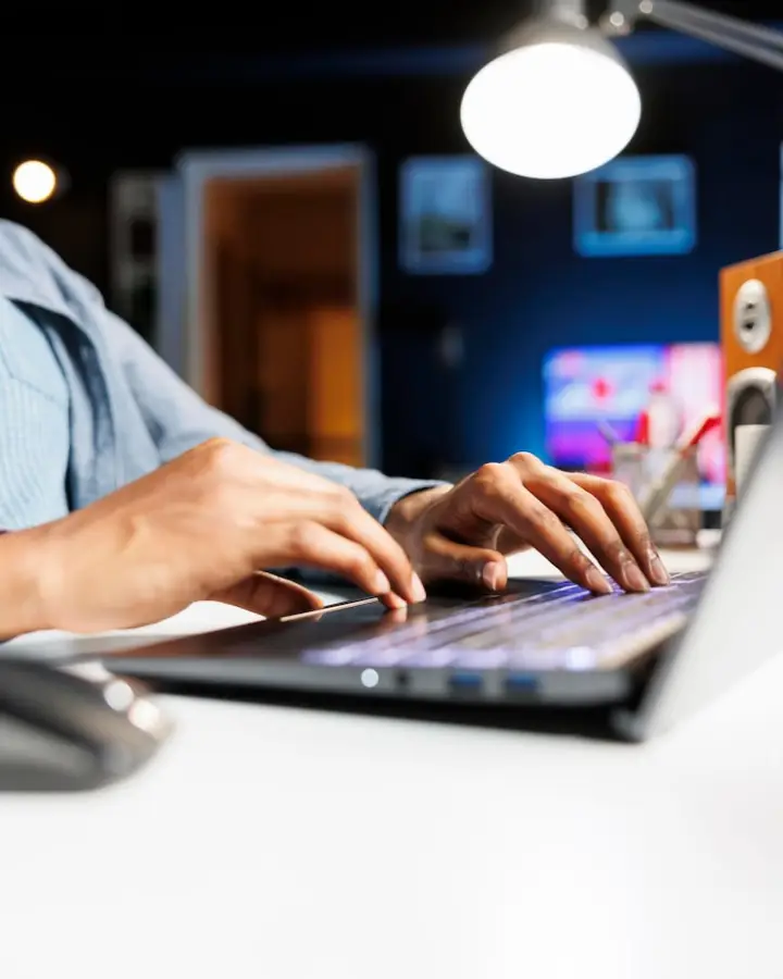Person typing on a laptop in a dimly-lit room, focusing on a desk lamp and hands on the keyboard.