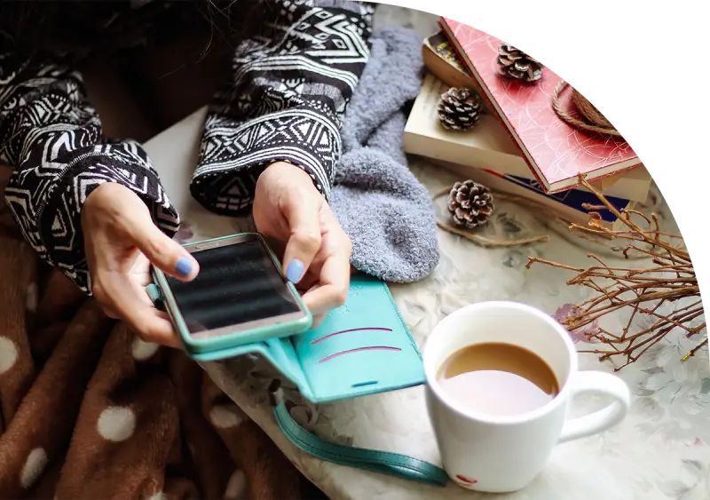 Hands holding a smartphone near coffee, books, and pine cones on a cozy table setting.